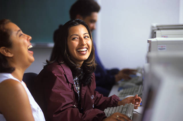 Three people sitting at computers side-by-side, the person in the middle is laughing.